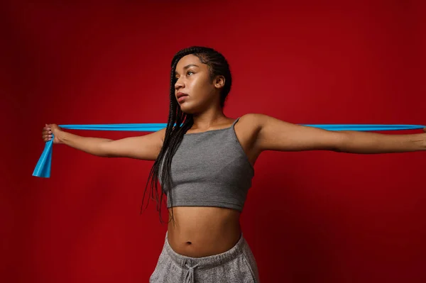 Determined young African woman with stylish dreadlocks stretching her arms using elastic rubber fitness band during routine workout against colored background with copy space