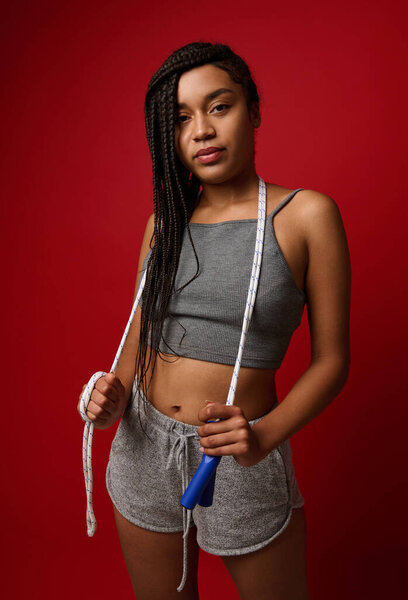Confident African woman with stylish dreadlocks and fit body posing against red background with jumping rope. Cardio workout, weight loss and fitness training concept with copy space for advertisement