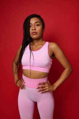 Kendine güveni tam, zarif rastaları olan, pembe spor giysileri giyen, kolları belinde kırmızı renkli bir arka plana yaslanmış, reklam için fotokopi alanı olan güzel bir Afrikalı kadın.