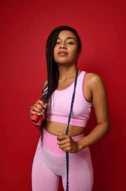 Pembe spor giyimli, kendine güvenen Afrikalı genç bir kadın boynunda ip atlarken poz veriyor, kameraya bakıyor, kırmızı arka planda fotokopi alanı ile izole edilmiş. Vücut pozitifliği, spor, spor konsepti