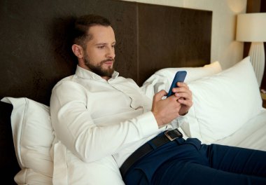 Yakışıklı Avrupalı genç sakallı adam, başarılı bir iş adamı cep telefonuyla geziniyor ve sohbet ediyor, bir otelin yatak odasında uzanıyor, iş gününden ve iş toplantılarından sonra dinleniyor.