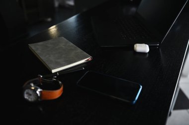 Ofis aksesuarları, kırtasiye malzemeleri, dizüstü bilgisayarlar, kutudaki kulaklıklar, saat, cep telefonu ve ahşap masadaki gri not defteri.