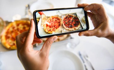 Elleriniz cep telefonuyla beyaz masa örtüsü ile servis edilen taze pişmiş lezzetli pizzanın resmini çekiyorsunuz. Cep telefonu canlı kipte