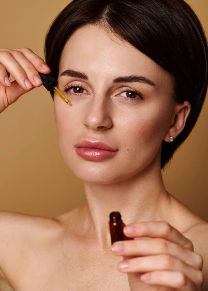 Güzel, sağlıklı, taze bir cilde sahip, elinde bir şişe kozmetik ürün ve kameraya bakarken yüzüne serum damlatan bir pipet tutan çekici esmer kadının yakın çekimi..