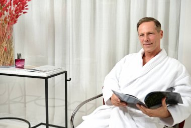 Beyaz bornoz giymiş yakışıklı, sağlıklı yaşam merkezinin özel odasında rahatlarken kameraya kendinden emin bir şekilde bakan, elinde bir dergi ile bir koltukta oturan,