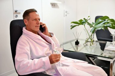 Yakışıklı, kendine güveni tam, elinde su olan beyaz bir adamın portresi. Sağlık spa salonunda koltukta oturuyor ve cep telefonuyla konuşuyor.