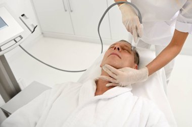 Estetikçi olgun bir adam için yüz gerdirme prosedürü uyguluyor. SPA 'daki modern ultrasonik ekipmanları deri dökülmesi, uyarılma ve su kaybı için kullanıyor. Erkeklerin estetik kozmetolojisi ve yaşlanma karşıtı cilt bakımı