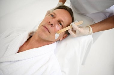 Estetik uzmanının yakışıklı, olgun Avrupalı bir adamın yüzüne tahta spatulayla nemlendirici yüz maskesi uygulaması. Erkeklerin güzellik tedavisi, cilt bakımı ve sağlık merkezindeki erkek kozmetolojisi konsepti