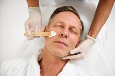 Güzellik uzmanının yakışıklı, olgun Avrupalı erkeklerin yüzüne nemlendirici yüz maskesi ve tahta spatula sürüşü. Sağlık kaplıcasında erkeksi güzellik, cilt bakımı ve erkek kozmetolojisi kavramı