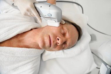 Estezi uzmanının, sağlıklı spa merkezinde modern ultrasonik medikal aparatlar kullanan olgun Avrupalı bir adamın yüzüne yüz gerdirme ve gençleşme karşıtı güzellik tedavisi uygulaması yapması.