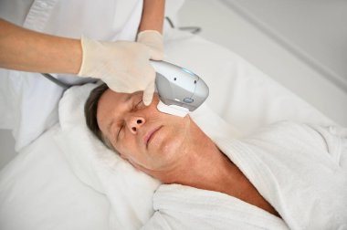 Yetişkin beyaz bir adamın sağlık merkezinden erkeksi bir cilt bakımı alırken güzellik uzmanının ultrasonik tıbbi cihaz kullanarak yüzüne yaşlanma karşıtı güzellik tedavisi uygulaması yapması genel bir bakış açısı.