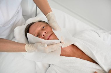 Güzellik estetisyeni, modern lüks bir spa ve sağlık merkezindeki masaj masasında dinlenen olgun ve çekici bir adamın cildine bakmak için yüz temizleme ve güzellik tedavileri uyguluyor.