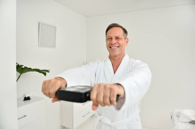 Gülümseyen, olgun, beyaz bornozlu, 50 yaşında beyaz bir beyaz adam, sağlık merkezinde biyolojik yaşını ve canlılığını belirlemek için tıbbi bir tartı ile tartılır.