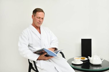 Beyaz bornozlu yakışıklı, çekici, olgun, beyaz beyaz beyaz adam koltukta oturup dergi okuyor, lüks sağlık merkezinin özel odasında dinleniyor.