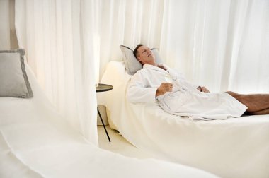 Beyaz Terry bornozlu rahat, yakışıklı, olgun beyaz adam elinde suyla bir bardak tutuyor ve lüks sağlık merkezindeki özel masaj odasındaki bir yatağa uzanıyor.