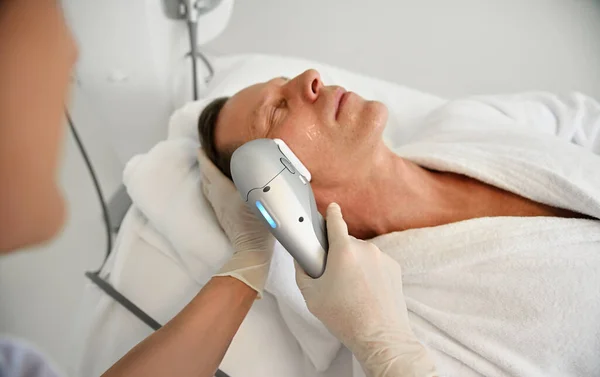 Close-up of a mature man relaxing during beauty procedures at wellness spa centre while aesthetician doing face lifting anti-aging beauty treatment on his face using ultrasonic medical apparatus