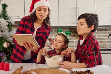 Kareli giysiler içinde mutlu bir aile, Noel Baba şapkalı güzel bir anne ve iki sevimli çocuğu, erkek ve kız birlikte Noel ekmeği ve kurabiyeleri hazırlıyorlar.