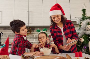 Kareli giysiler içinde mutlu bir aile, Noel Baba şapkalı güzel bir anne ve iki sevimli çocuk, erkek ve kız birlikte Noel ekmeği ve kurabiyeleri hazırlıyorlar.