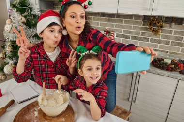 Neşeli, güzel İspanyol anne boynuz çemberiyle sevimli çocukları ve Noel Baba şapkasıyla birlikte eğleniyor, Noel ekmeği pişiriyor ve akıllı telefondan web kamerasına bakarak selfie çekiyor.
