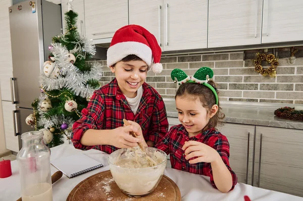 Mutlu Noeller ve mutlu çocukluk kavramları. Elf çemberli şirin küçük kız ve ağabeyi Noel Baba şapkalı sevimli beyaz oğlan ev mutfağında fırın yapmaktan keyif alıyorlar.