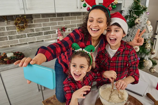 Ev mutfağında Noel havası. Sevinçli anne ve sevimli çocukları dişlek gülümseme ile akıllı telefonun web kamerasına bakıyor, selfie çekiyor, birlikte yemek pişirirken video kaydediyorlar.