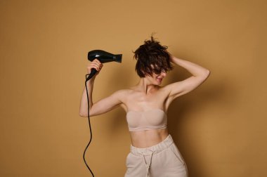 Bej arka planda, gözünün altında kolajen lekeleri olan çekici bir kadının, saç kurutma makinesinden ıslak saçına sıcak hava akışını yöneten ve onu kurutan, bej rengi arka planda izole edilmiş bir portresi.