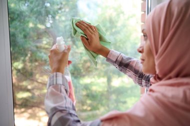 Yakın plan. Kafası tesettüre bulanmış temizlikçi. Pencere camını bez ve sprey deterjanla yıkıyor. Olumlu Arap kadın profesyonel ev temizliği yapıyor, evde hijyen hizmeti veriyor.