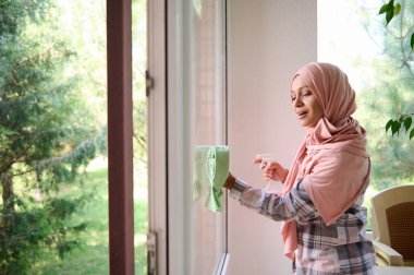 Kafası tesettüre bulanmış güzel bir Arap kadının yan portresi. Pencereleri siliyor, deterjan sıkıyor, süngerle siliyor, evde bahar temizliğinin keyfini çıkarıyor.