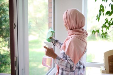 Kafası tesettüre bulanmış Arap Müslüman bir kadın pencereleri siliyor, lekeleri temizliyor, deterjan sıkıyor ve arka planda bahçeli evin genel bahar temizliğinin keyfini çıkarıyor.