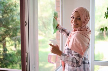 Başı tesettüre bulanmış, keyifli, dişlek gülümseyen, omzunun üzerinden kameraya bakan, pencereleri yıkayan, evin genel bahar temizliğinin keyfini çıkaran, keyifli bir Arap kadını.