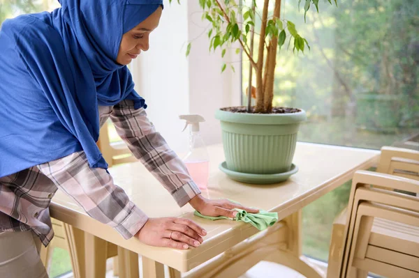 Kafası tesettüre bulanmış orta yaşlı bir kadın evde angarya işler yapıyor. Oda servisi konsepti. Arap Müslüman kadın verandada mikrofiber bez kullanarak tozdan masayı temizlemeye ve silmeye odaklanmış..
