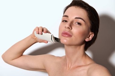 Ultrasonik deri soyma işlemi sırasında çekici, doğal güzellikte bir kadının yüzüne yakın çekim. Yüz derisi temizleniyor, siyah noktalar siliniyor, beyaz arkaplanda kopya reklam alanı ile izole ediliyor