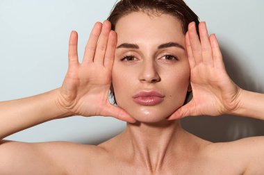 Yakın plan. Genç, zarif, beyaz bir kadının, sağlıklı, parlayan yüzüne elleriyle dokunup kameraya baktığı, kaldırma ve kaplıca tedavisi için yumuşak masaj hareketleri yaptığı bir vesikalık fotoğrafı.