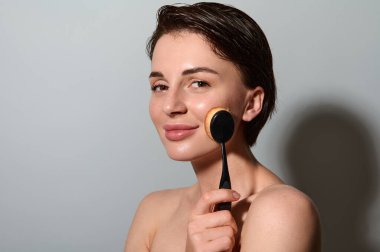 Çekici beyaz tenli, sağlıklı ve doğal makyajlı bir esmer, makyaj fırçasıyla bir vakıf kuruyor, kameraya gülümsüyor, gri arka planda fotokopi alanı ile poz veriyor.