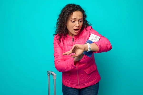 Stunned Hispanic woman in bright pink jacket looks at her watch with surprise and fear, being late for her flight, holding passport with air ticket, standing next to a suitcase against blue background
