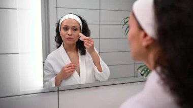 Güzel, siyah saçlı, beyaz waffle bornozlu güzel bir kadın. Yeşim taşı Gua-Sha masaj aleti kullanarak yüz masajı yapıyor. Banyodaki ayna yansımasına bakıyor. Yaşlanma karşıtı, cilt bakımı konsepti