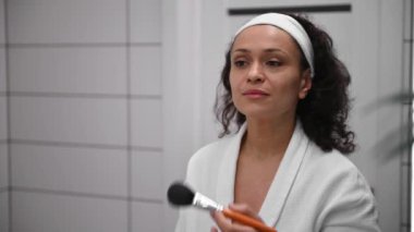 Beyaz bornozlu güzel bir kadın banyo aynasının önünde duruyor ve makyaj fırçasıyla yüzüne pudra sürüyor, dişlek bir gülümsemeyle ayna görüntüsüne bakıyor.