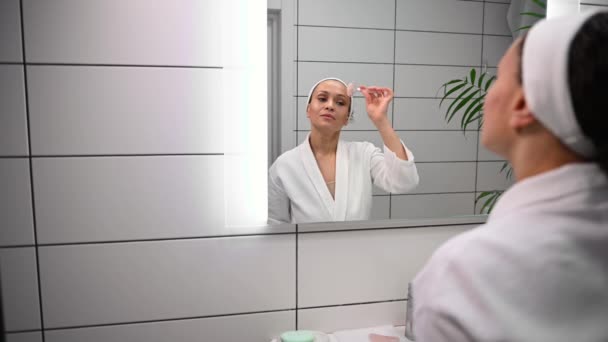 バスルームで彼女の鏡の反射を見て 玉石ローラーマッサージを使用して顔のマッサージを行う白いワッフルバスローブの魅力的な暗い髪のきれいな女性 アンチエイジング スキンケアのコンセプト — ストック動画