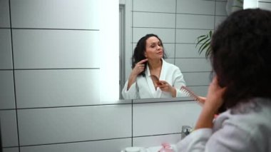 Güzel, orta yaşlı, kıvırcık, siyah saçlı, beyaz waffle bornoz giyen bir kadın, banyo aynasının önünde duruyor ve saçlarını tarıyor, yansımasına bakıyor. Hijyen, vücut bakımı kavramları.
