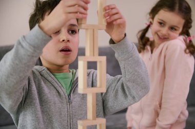 Yakın plan. Yakışıklı okul çocuğu, ahşap bloklardan karmaşık uzun yapılar inşa etmeye odaklanmış, neşeli küçük kız kardeşinin arka planına karşı, tatlı tatlı gülümsüyor, yapısına bakıyor