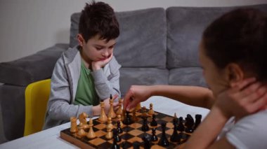 Konsantre olmuş beyaz okul çocuğu annesiyle oynarken satranç hareketlerini düşünerek satranç stratejisi geliştiriyor. Mantık geliştirme, boş masa oyunları, eğlence ve eğitim kavramı