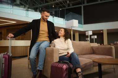 Woman and man in casual clothes with suitcases sitting on armchair in the international airport terminal lounge, looking at each other, discussing forthcoming flight, waiting for airplane departure