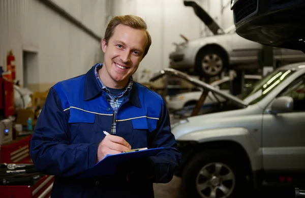 汽车维修 汽车保修服务的概念 汽车技师写在剪贴板上的一个车间 年轻的高加索技师笑着看着相机 为车库里的汽车修理做检查清单 — 图库照片