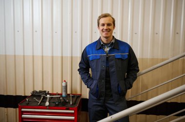 Yakışıklı beyaz bir adamın portresi, garaj tamircisi, teknisyen kameraya bakıp gülümseyen bir kutunun yanında duran iş aletleriyle otomobilleri ve araba bakım istasyonunu incelemek ve tamir etmek için.