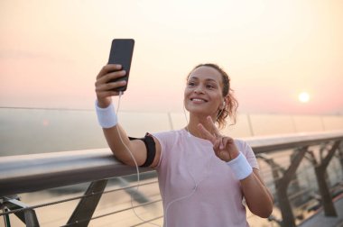 Çekici bir kadın neşeli bir gülümsemeyle gülümser ve kameraya bakarak barış işareti gösterir. Elinde akıllı bir telefonla, güneş doğduktan sonra bir şehir köprüsünde antremandan sonra selfie çekerken.