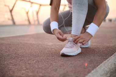 Tanınmaz haldeki spor ayakkabısı bağlayan, sabah koşusuna hazırlanan ve şafak vakti şehir köprüsündeki koşu bandında spor yapan bir kadın resmi.