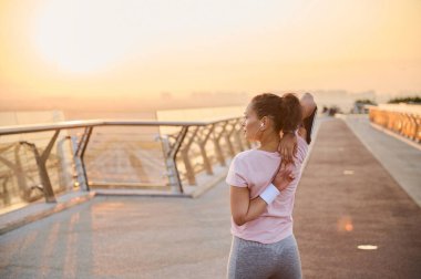 Kararlı bir bayan atlet, sporcu kadın kollarını uzatıyor, kaldırıyor, gün doğumunda şehir köprüsünde egzersiz yapıyor. Spor, sağlık, vücut bakımı, spor ve aktif yaşam tarzı kavramı