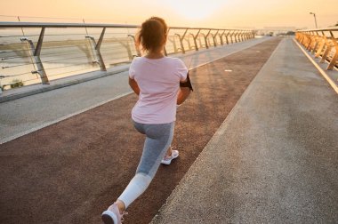 Spor kıyafetli bayan bir sporcunun bacak kaslarını esnetirken, hamleler yaparken, sabahın erken saatlerinde şehir köprüsünde spor yaparken görüntüsü. Genç sporcu kadın şafakta dışarıda egzersiz yapıyor..
