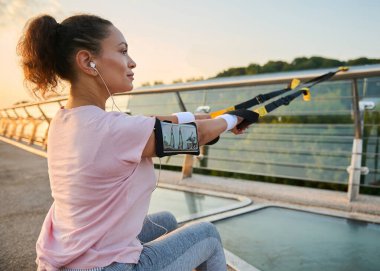 Yazın şehir ortamında yoğun süspansiyon eğitimi sırasında kayışlarla egzersiz yapan yakın plan atletik kadın. Spor giyimli kadın sporcu, süspansiyon ipleri çekiyor, çömeliyor, hamlesini yapıyor.