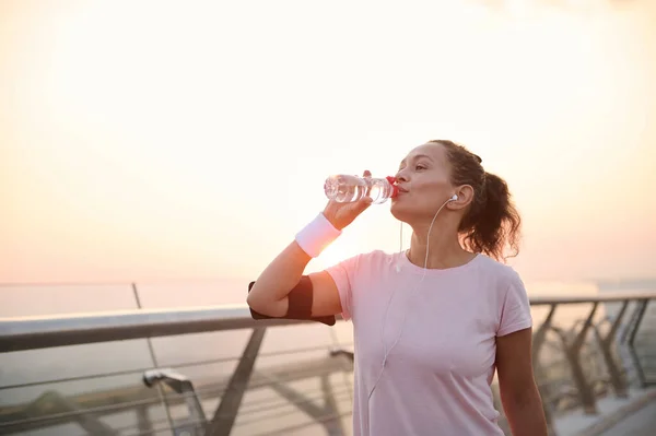 Happy determined athlete, beautiful Hispanic woman wearing pink t-shirt, smartphone holder and terry wristbands stands on a city bridge and drinks water, resting after early morning jog at sunrise